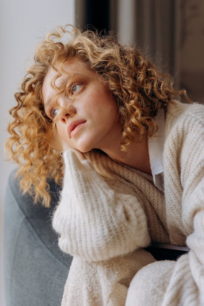 A woman with curly hair gazes thoughtfully while sitting indoors, wrapped in a cozy knit sweater.