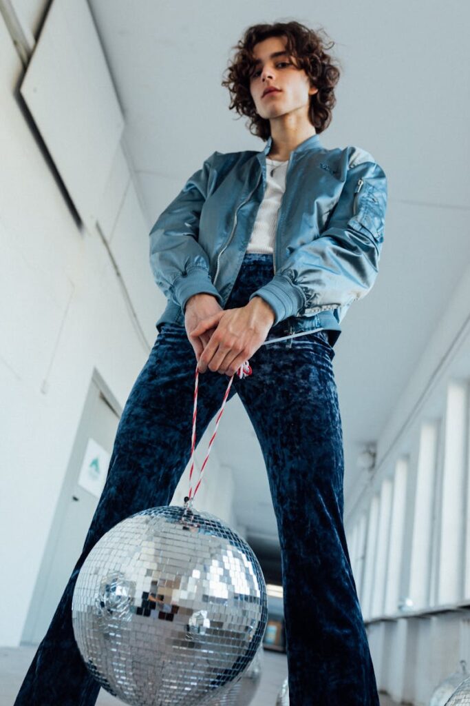 Young adult with curly hair stylishly posing with a disco ball in a modern hallway. Low angle shot.