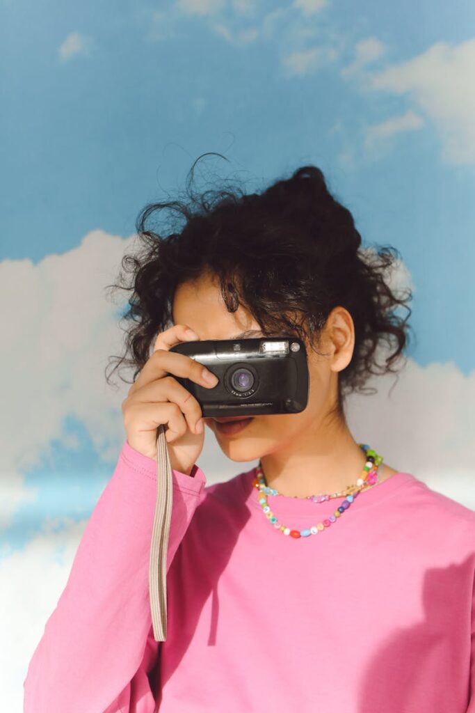 Portrait of a young woman with curly hair capturing a photo with an analog camera against a cloud-filled sky.