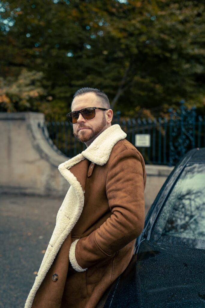Confident man in a brown coat leaning against a car, exuding style and sophistication.
