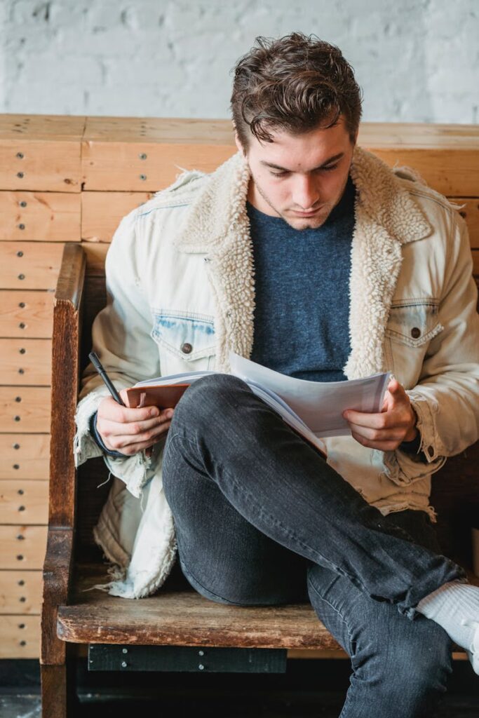 A young man attentively reads papers while sitting on a wooden bench indoors.