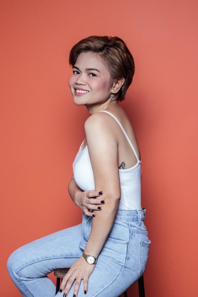 Young woman in white tank top and jeans posing and smiling against orange-red backdrop.