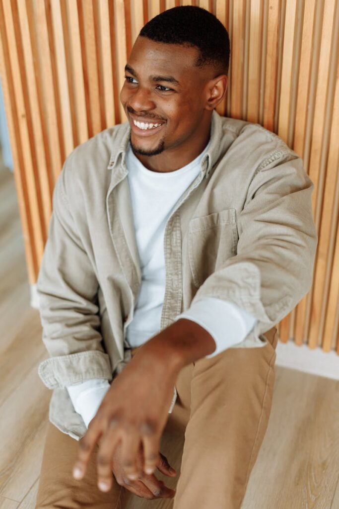A fashionable man in casual clothing smiling while seated against a wooden backdrop.