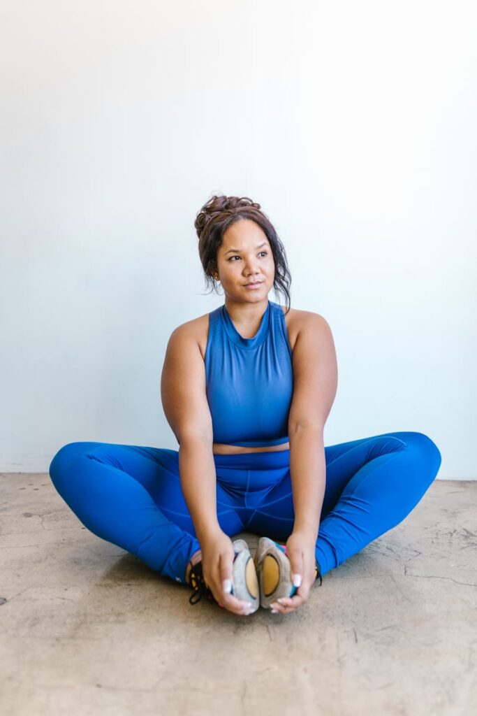 Plus-size woman in blue activewear sitting on floor and stretching indoors.