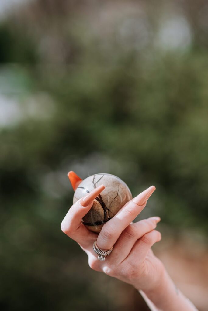 Unrecognizable female with manicure showing small brown round shaped mineral in hand while standing on street with green trees on blurred background