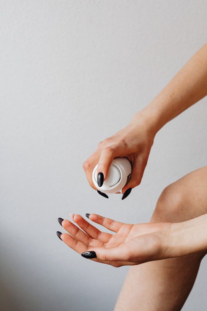 Close-up of a woman's hands with black nail polish applying skincare cream against a light background.