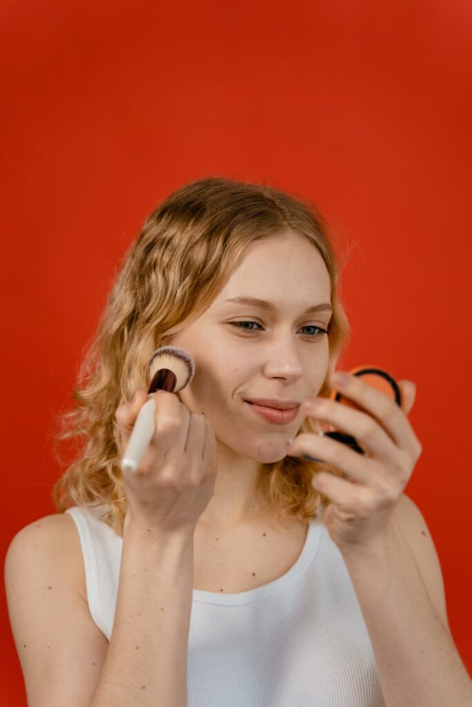Portrait of a smiling woman applying makeup with a blush brush and mirror against a red background.
