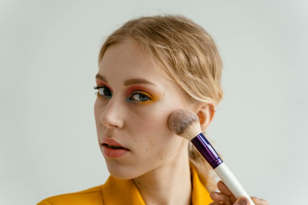 A woman applying blush with a makeup brush, showcasing a colorful eyeshadow look.