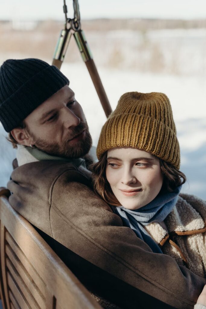 A couple in warm jackets and beanies enjoys a romantic moment on a wooden swing in winter.