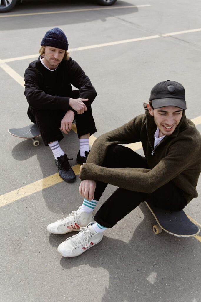 Casually dressed young men sitting on skateboards in a sunny parking lot setting.