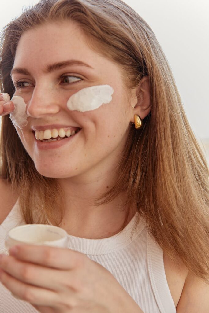 A smiling woman applies skincare cream to her face indoors, highlighting a beauty routine.