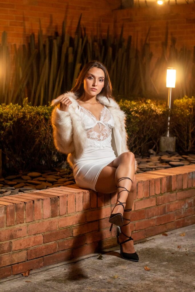 A stylish woman in a white dress and fur jacket poses gracefully outdoors at night.