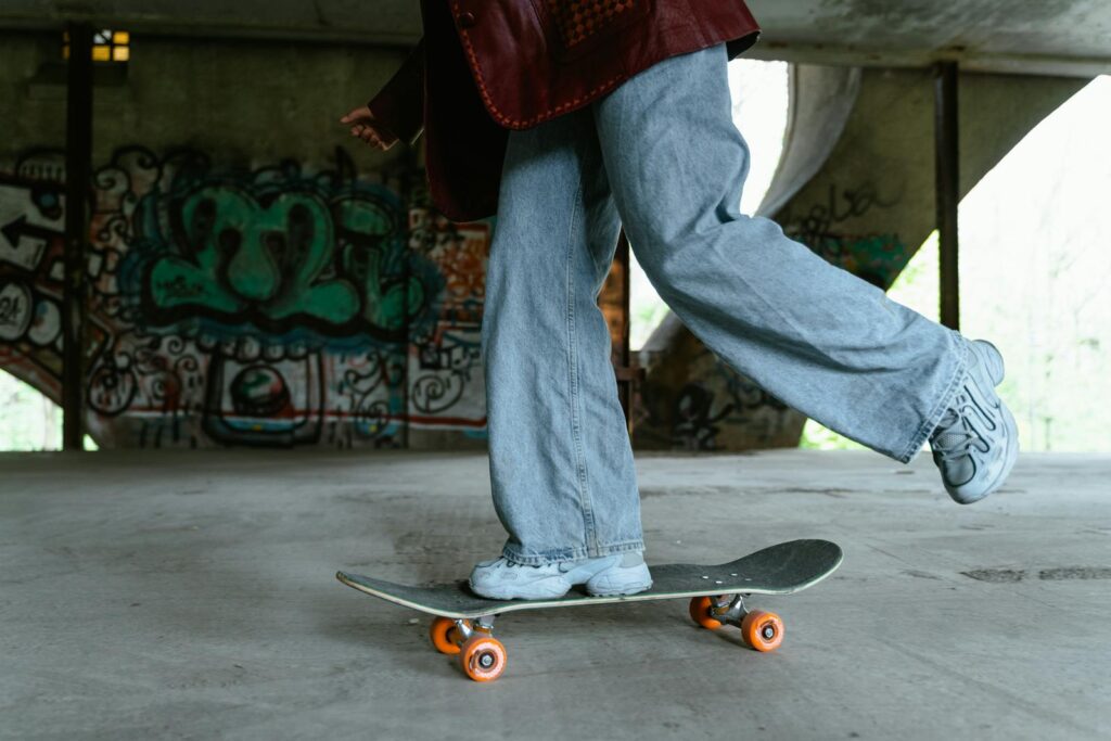 A skateboarder in denim jeans skates over a concrete floor in an urban area with graffiti.