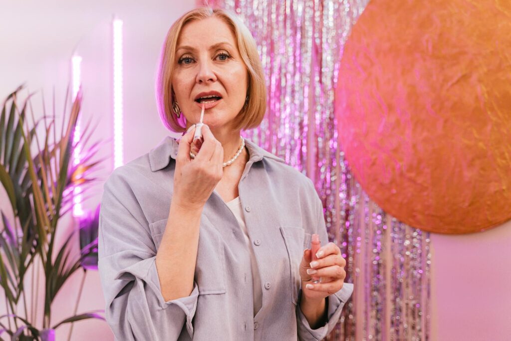 A mature Caucasian woman applies lipstick in a stylish, colorful setting. Great for beauty and lifestyle themes.