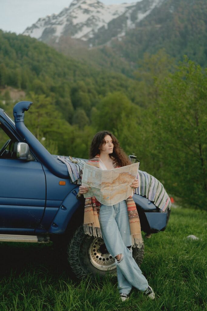 A woman leans on a blue car in a mountain scene, reading a large map outdoors.