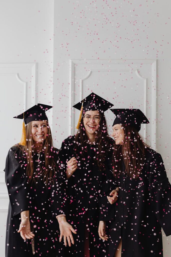 Three women graduates joyfully celebrate with confetti indoors, showcasing excitement and success.