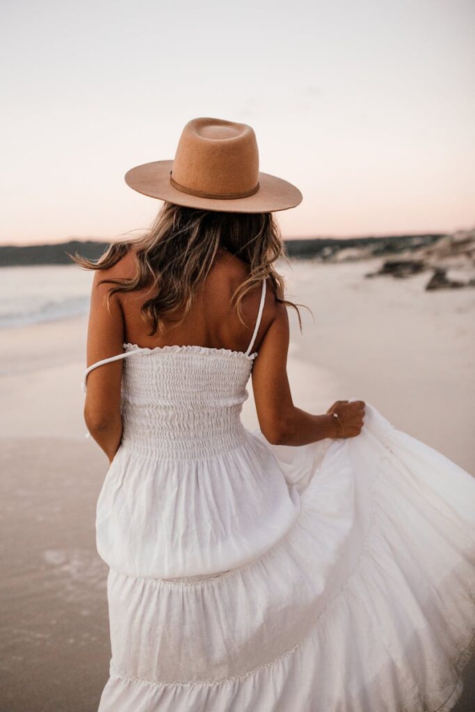 A woman in a white dress and hat walks along a serene beach at sunset.