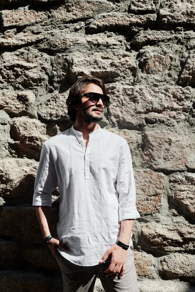 Bearded man in white shirt and sunglasses posing against rock wall.