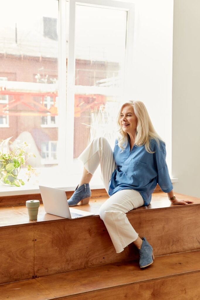 Elderly woman enjoying a relaxing moment with laptop by a bright window.