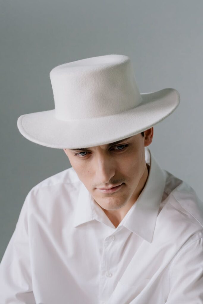 Stylish portrait of a man wearing a white fedora and shirt, with a soft neutral background.