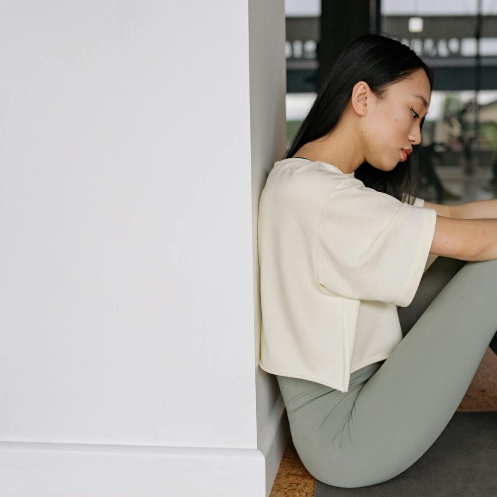 Side view of a young woman in yoga attire relaxing indoors, sitting against a wall.