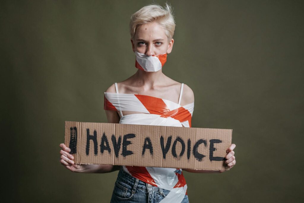 Woman wrapped in tape holding a protest sign 'I Have a Voice' in a studio setting.