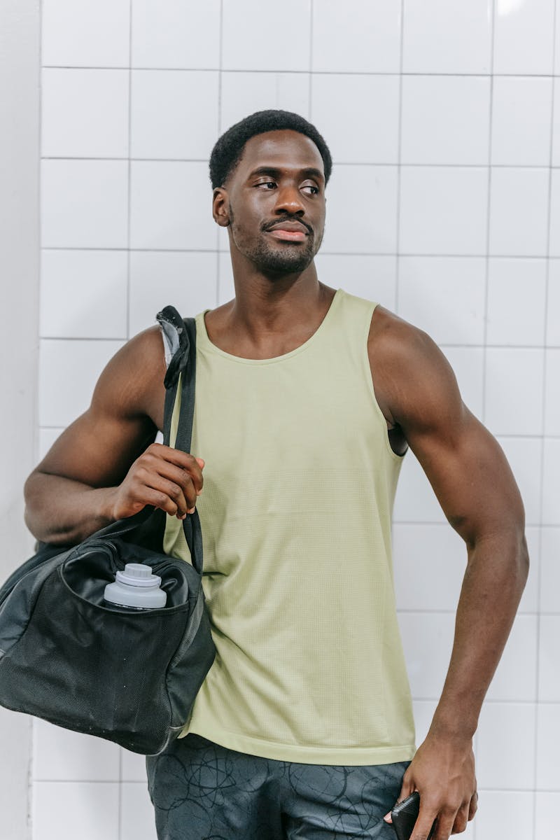 Athletic man in tank top holding a sports bag inside a gym locker room, ready for workout.