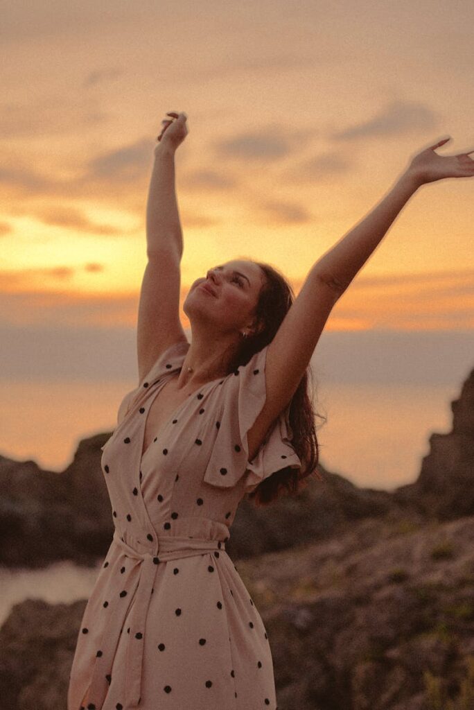 A woman in a polka dot dress expresses freedom and joy by the sea at sunset.