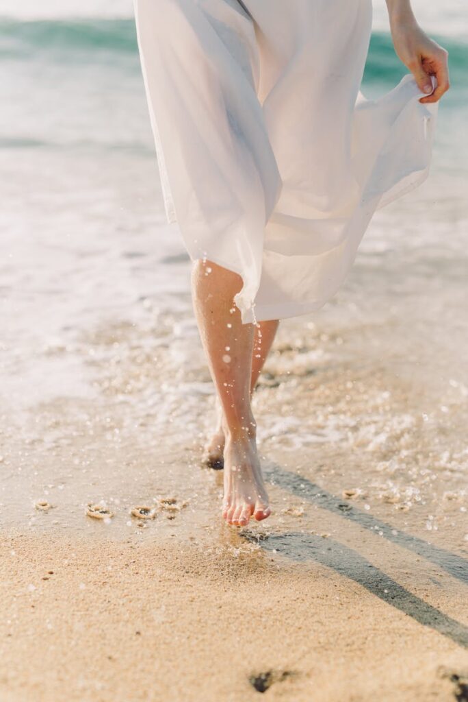 A woman in a white dress walks barefoot along a sunlit beach with gentle ocean waves.