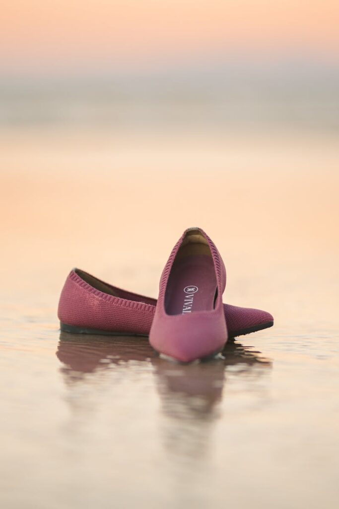 Elegant pink ballet flats resting on wet sand during a serene sunset.