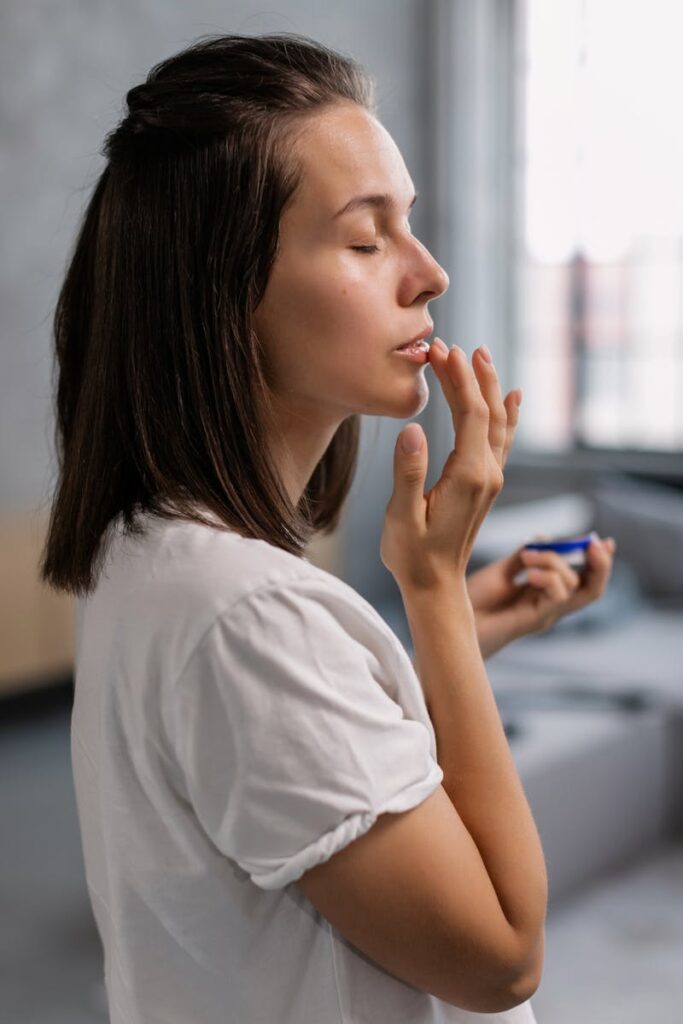 A side view of a woman applying lip balm with eyes closed, creating a serene moment indoors.