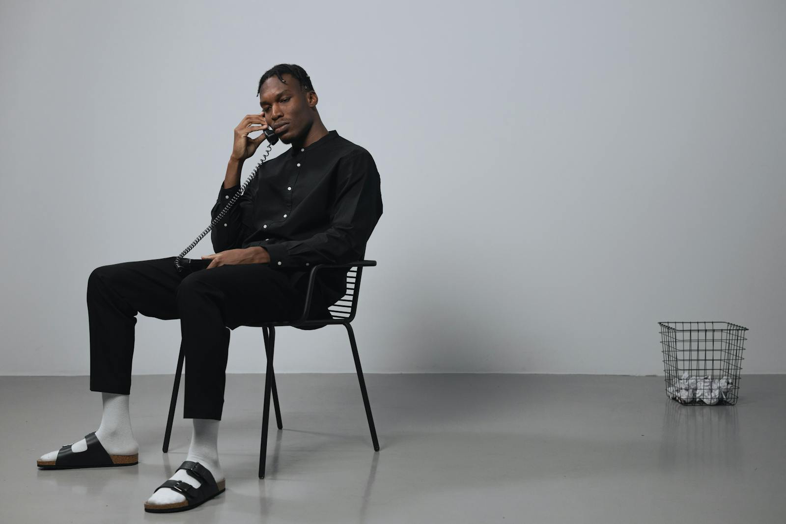 African American man in black attire talking on a phone while sitting indoors with a minimalistic setup.