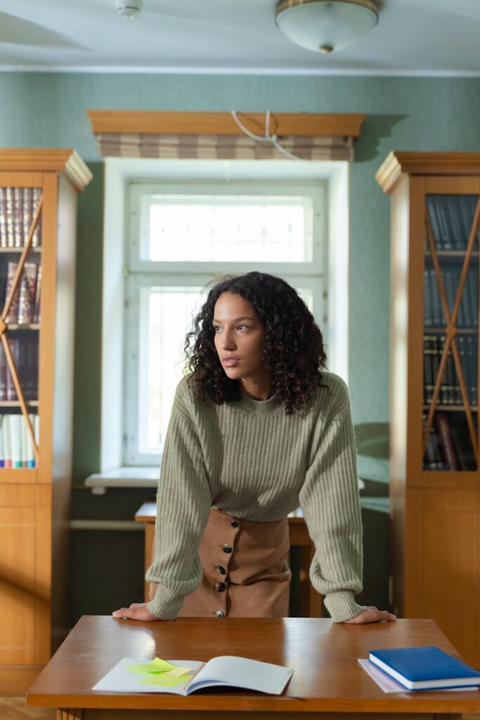A stylish woman in a library leans on a desk, exuding confidence and intellect.