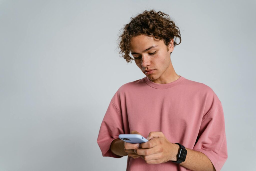 Teenager with curly hair using a smartphone indoors, wearing a pink t-shirt.