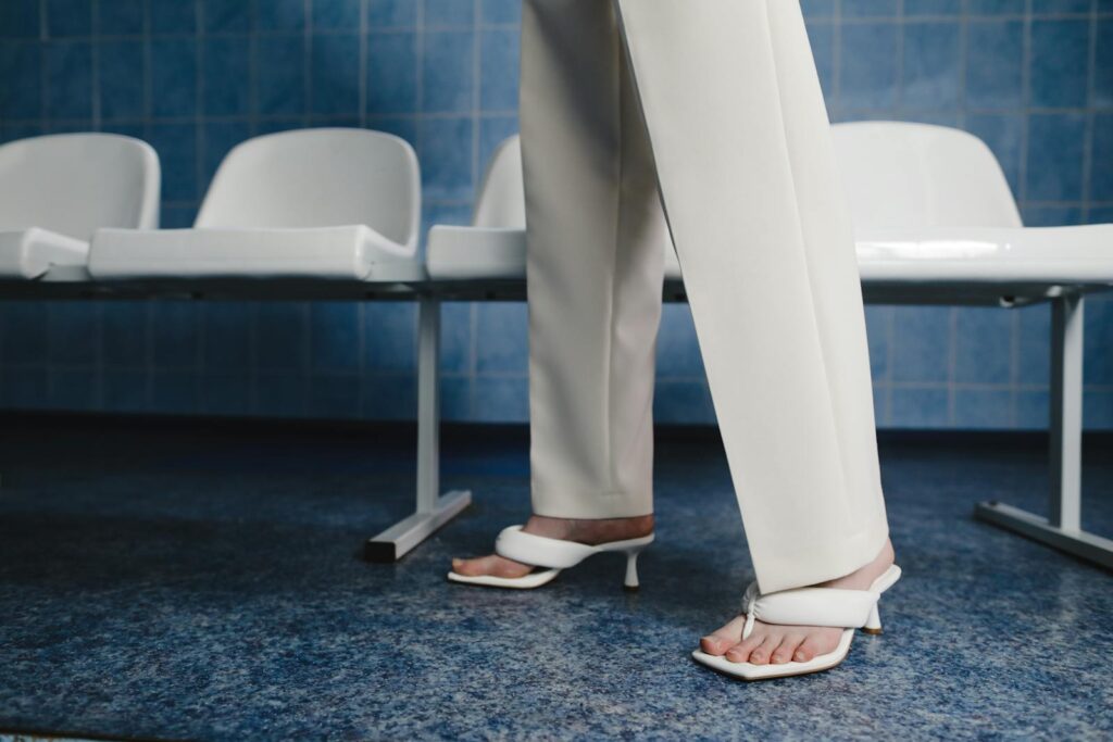 Minimalist fashion image of a woman in white pants and sandals standing indoors by seats.