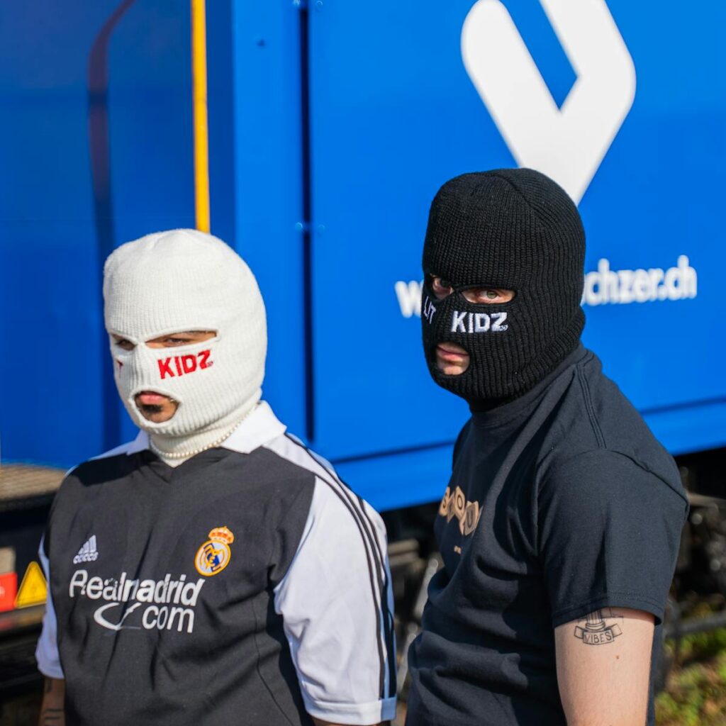 Two masked individuals in branded shirts pose outdoors against a blue background.