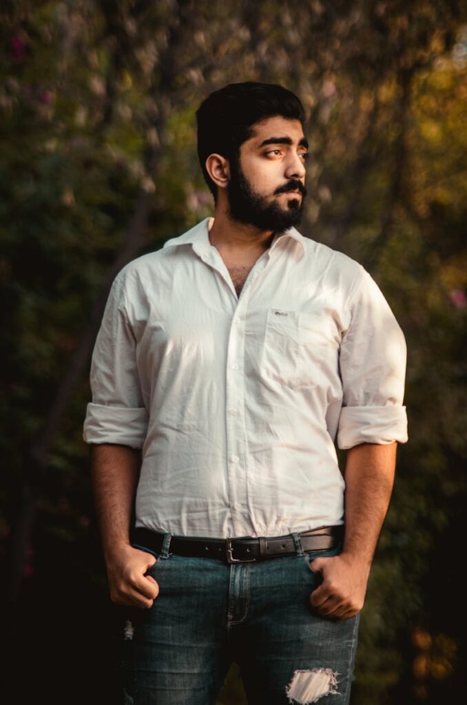 Portrait of a bearded man standing outdoors, wearing a white shirt, with hands in pockets, in soft natural light.