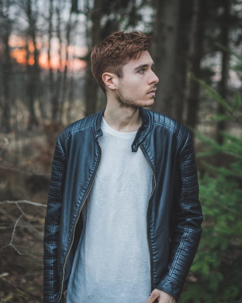 Young man in a black leather jacket standing in a forest during twilight, styled fashion portrait.