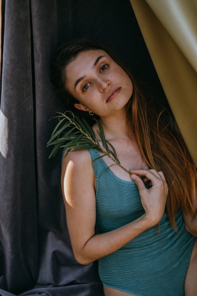 Beautiful woman in a turquoise swimsuit holding leaves, posing indoors with natural lighting.