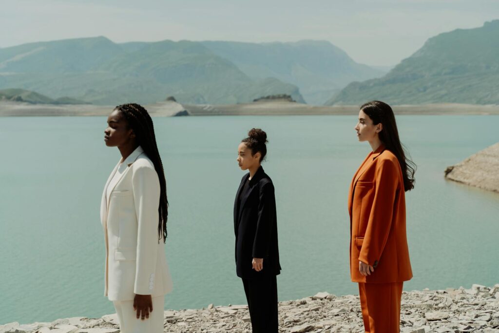 Three women in suits stand by a serene mountain lake, showcasing diversity and style.