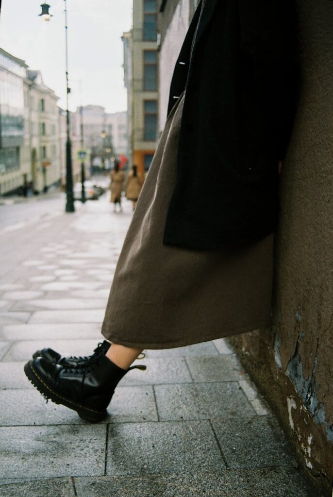 Person wearing black boots leaning against a wall on a wet, urban street, creating a moody city vibe.