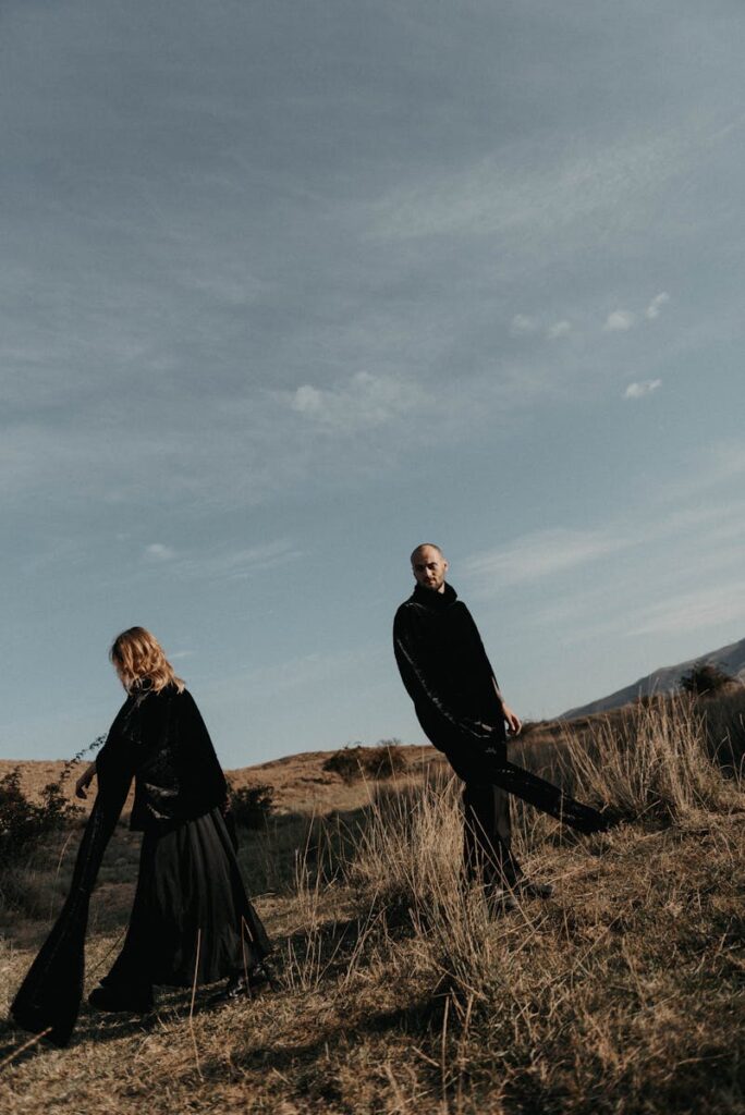 Man and woman in dark attire stand in a dry grassy outdoor landscape under a cloudy sky.