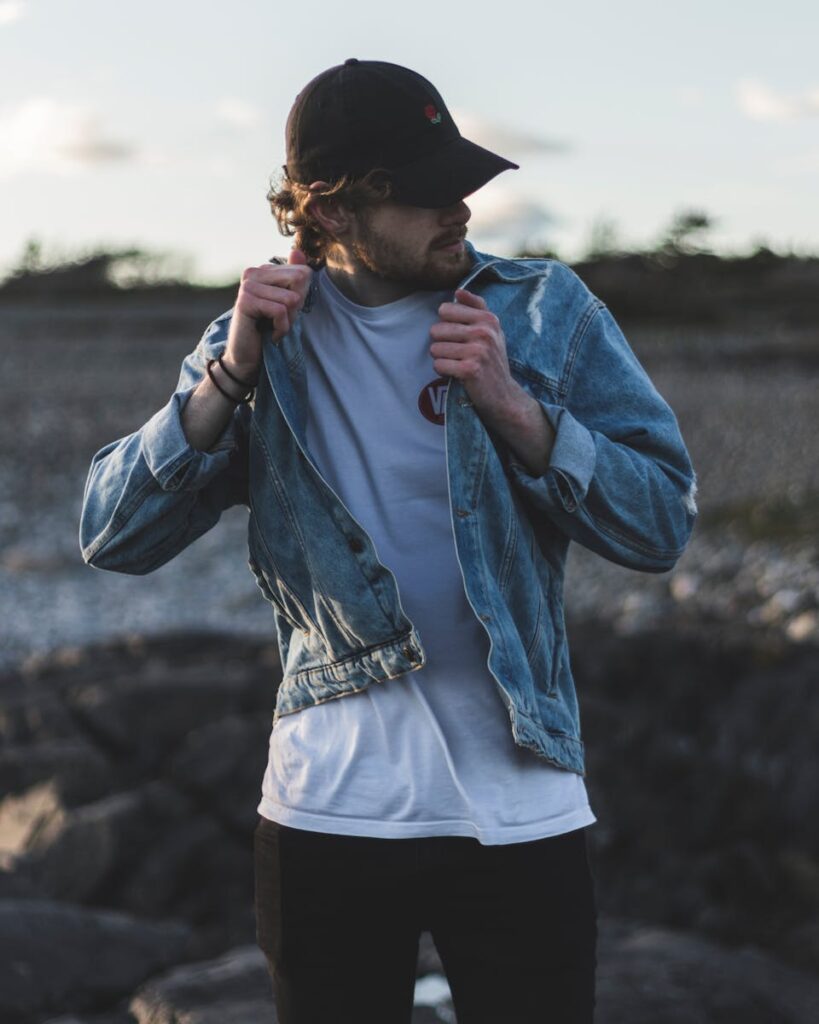 Man in a denim jacket and cap on a rocky outdoors setting, showcasing casual fashion.