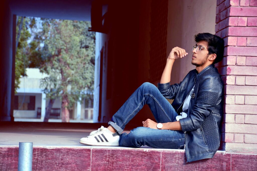 Stylish young man in denim and leather jacket sits thoughtfully outdoors.