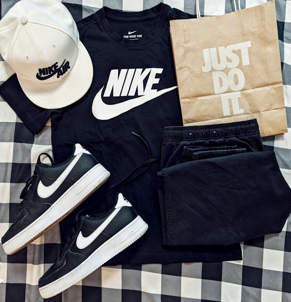 Flat lay of Nike apparel including t-shirt, cap, sneakers, and paper bag on checkered surface.