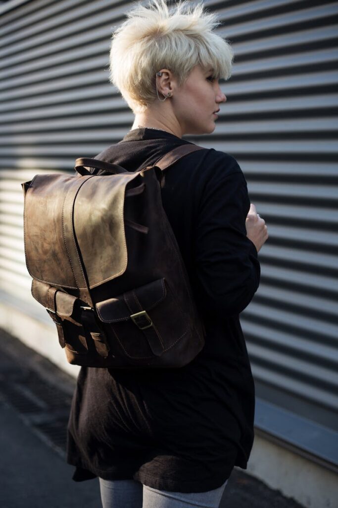 Side view of a stylish woman wearing a leather backpack against a corrugated metal wall.