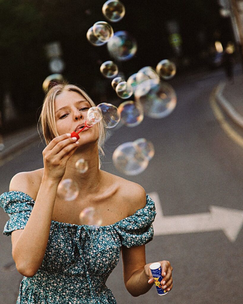 A blonde woman in a floral dress enjoys blowing bubbles while standing on a city street.