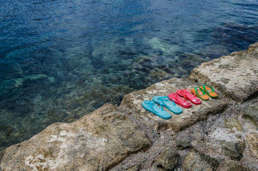 Vibrant flip flops on a rocky seashore beside clear ocean waters.