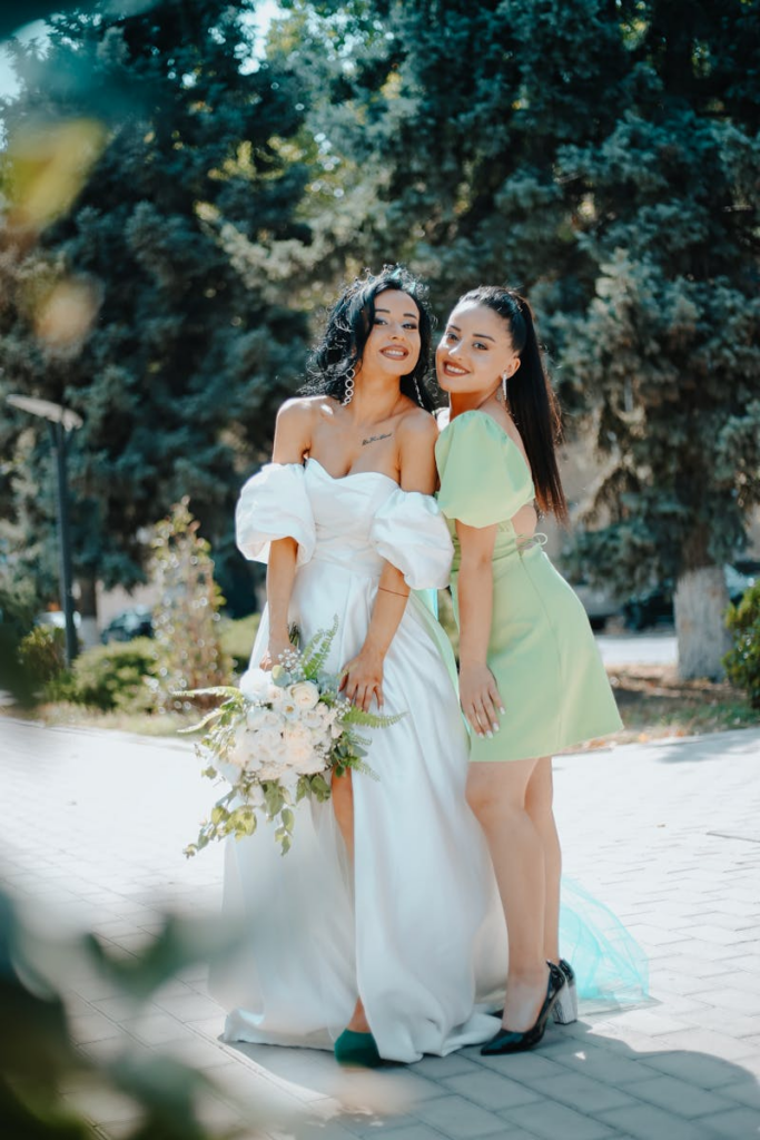 Bride and friend posing outdoors, vibrant summer day, elegant dresses.