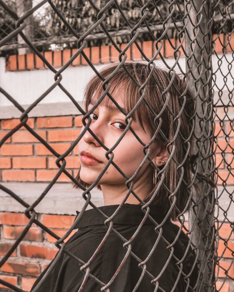 Portrait of a young woman standing behind a chain link fence with a brick wall background.
